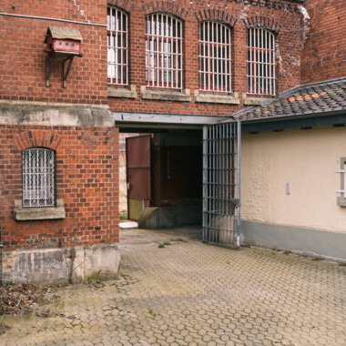 Ein Bild im Innenhof der JVA Rennelberg. Ein großes Tor in einer hohen Backsteinmauer, dahinter das ehemalige Gefängnisgelände. Über der Mauer verläuft Stacheldraht, im Hintergrund sind die umliegenden Wohnhäuser.